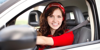 Ung kvinna med långt brunt hår och röd tröja sitter leende bakom ratten i en bil.