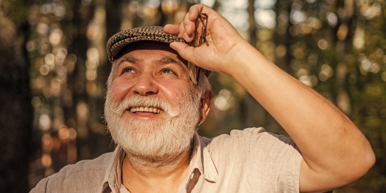 Leende äldre man med vitt skägg och keps står utomhus i soligt skogsparti.