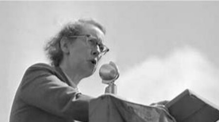 En äldre kvinna talar i mikrofon utomhus, klädd i kavaj, svartvit bild.