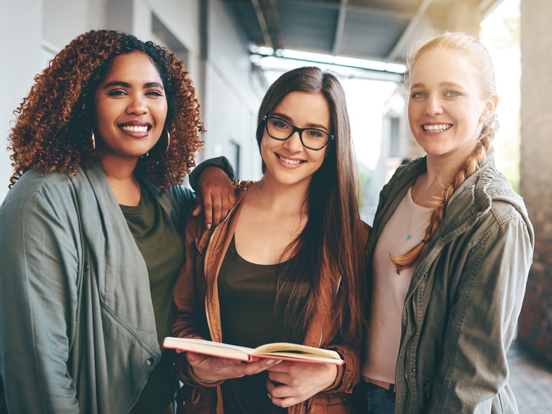 Tre unga kvinnor står tillsammans utomhus, leende och håller en bok.