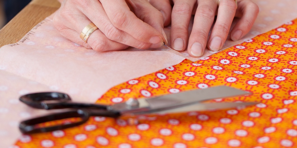 Händer som nålar fast mönsterpapper på orange tyg med sax och krita bredvid.