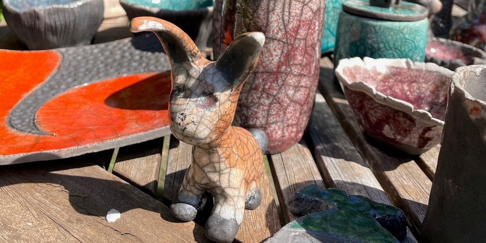 Keramikföremål, inklusive en kaninfigur, står på ett träbord utomhus med grönska i bakgrunden.