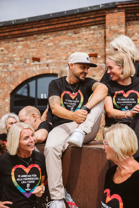 En grupp glada människor i svarta tröjor med regnbågshjärtan sitter utomhus vid en tegelvägg.