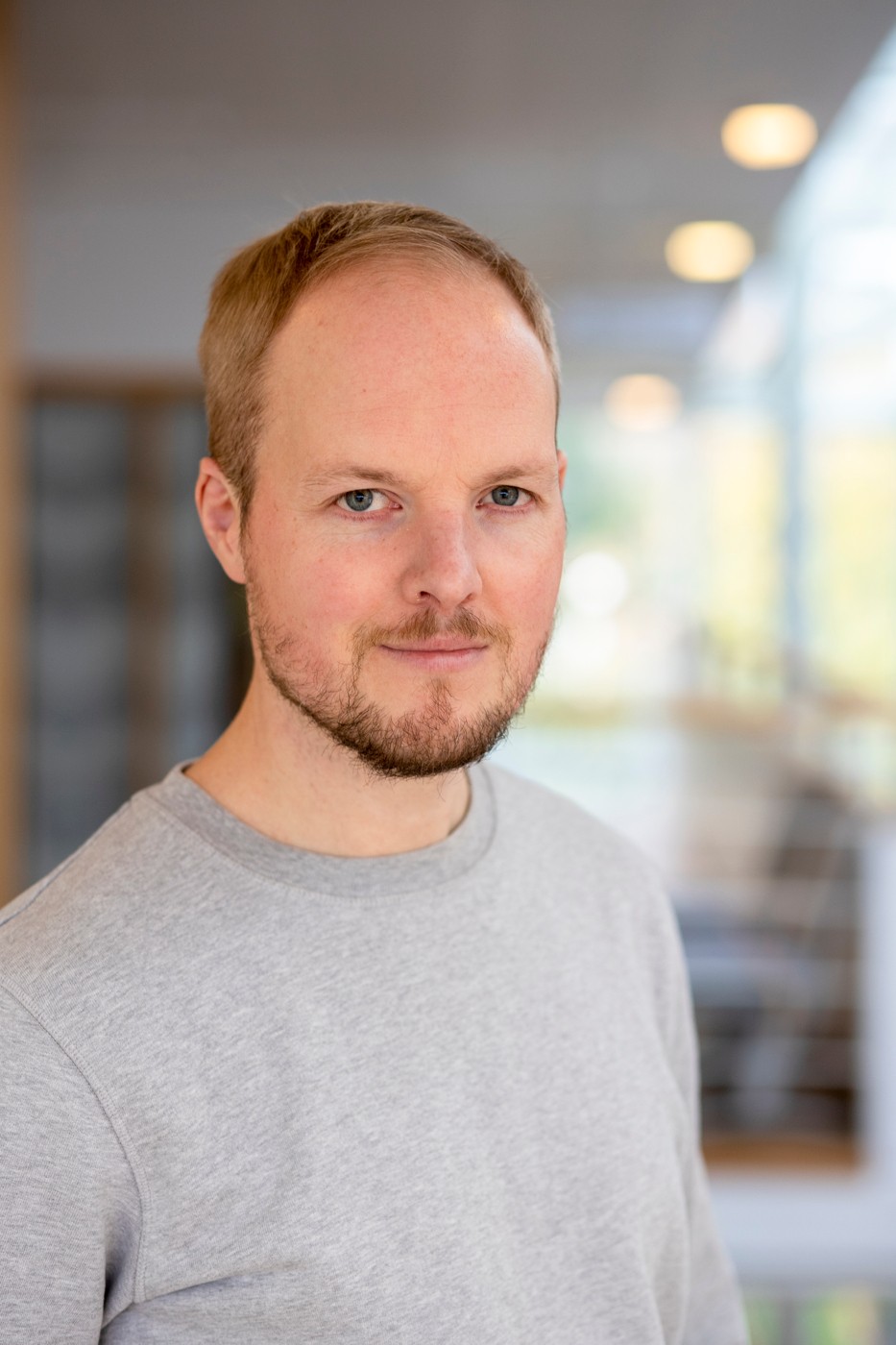 Man med kort hår och skägg bär en grå tröja och står inomhus i ett ljust, modernt rum.