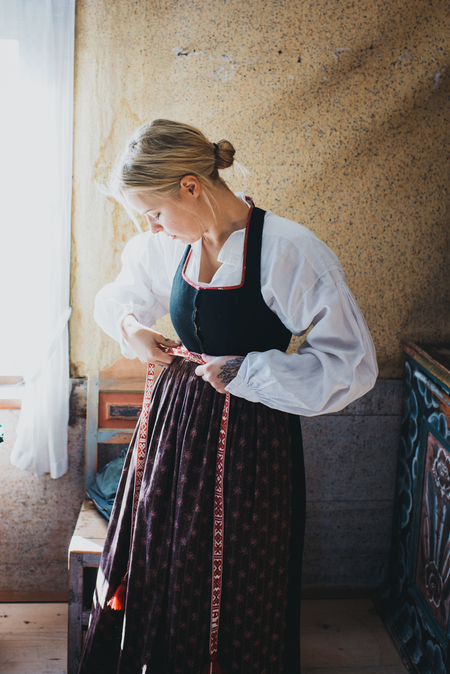 Kvinna klädd i traditionell folkdräkt knyter ett band runt midjan i ett solbelyst rum.