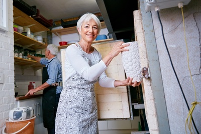 Äldre kvinna öppnar en keramikugn i en ljus verkstad, en man arbetar i bakgrunden.