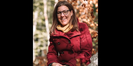 Kvinna med glasögon och röd kappa sitter utomhus bland höstlöv och ler mot kameran.
