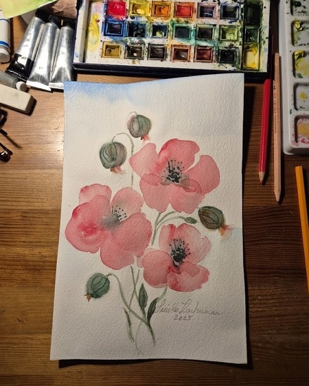 Akvarellmålning av rosa blommor på ett bord med färglåda, penslar och pennor runtom.