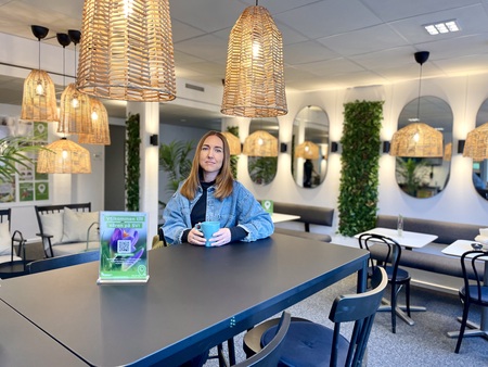 Kvinna sitter vid ett bord med en kopp i ett modernt, ljust café med speglar och växtdekorationer.