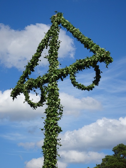 En midsommarstång klädd med gröna blad mot en blå himmel med vita moln.