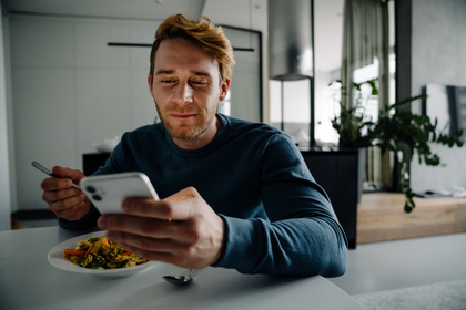 Man sitter vid ett bord, äter mat och tittar på sin mobiltelefon i ett modernt hem.