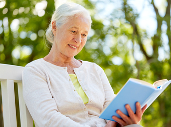 Äldre kvinna med vitt hår sitter utomhus på en bänk och läser en bok, omgiven av grönska.