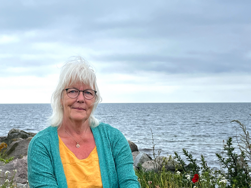Äldre kvinna med vitt hår och glasögon sitter vid havet, klädd i gul tröja och turkos kofta.