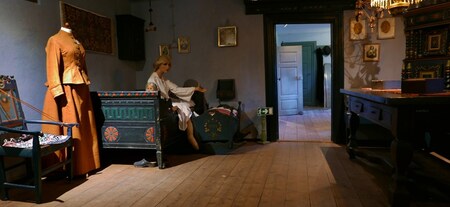 Ett historiskt rum med trägolv, gammaldags möbler, en skyltdocka i kläder och en sittande docka på en bänk.