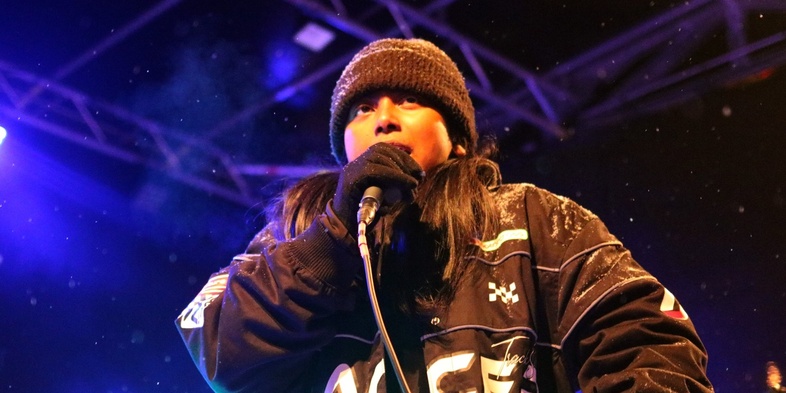 Person med mössa och mikrofon uppträder på en scen med färgglad belysning.