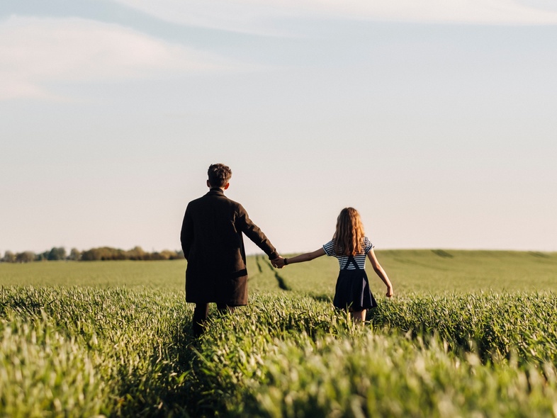 Två personer, en vuxen och ett barn, går hand i hand genom en grön åker under blå himmel.