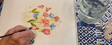 En person målar blommor med akvarellfärger vid ett bord fullt av konstmaterial.