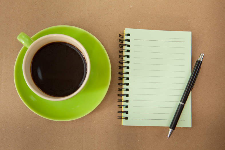 En kopp svart kaffe på ett grönt fat bredvid ett tomt spiralblock och en penna på ett brunt bord.
