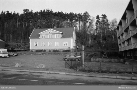 Svartvit bild på ett hus med sadeltak, en parkeringsplats och en bil, omgiven av skog och ett flerfamiljshus.