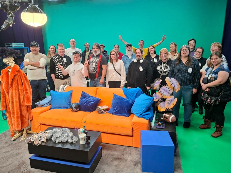 En grupp människor står i en TV-studio med grönskärm, orange soffa och rekvisita.