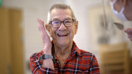 Leende äldre man i röd-rutig skjorta vinkar, inomhusmiljö, en person med munskydd syns delvis.