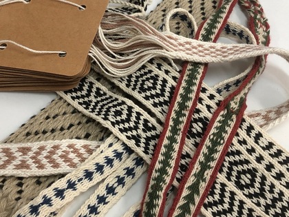 Flera handvävda band i olika mönster och färger ligger bredvid bruna pappersetiketter med snören.