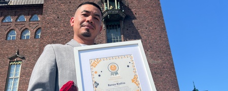 Man i grå kostym håller ett diplom och en röd ros framför en tegelbyggnad utomhus.