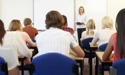 En lärare står framför en klass och undervisar elever som sitter vid bänkar i ett klassrum.