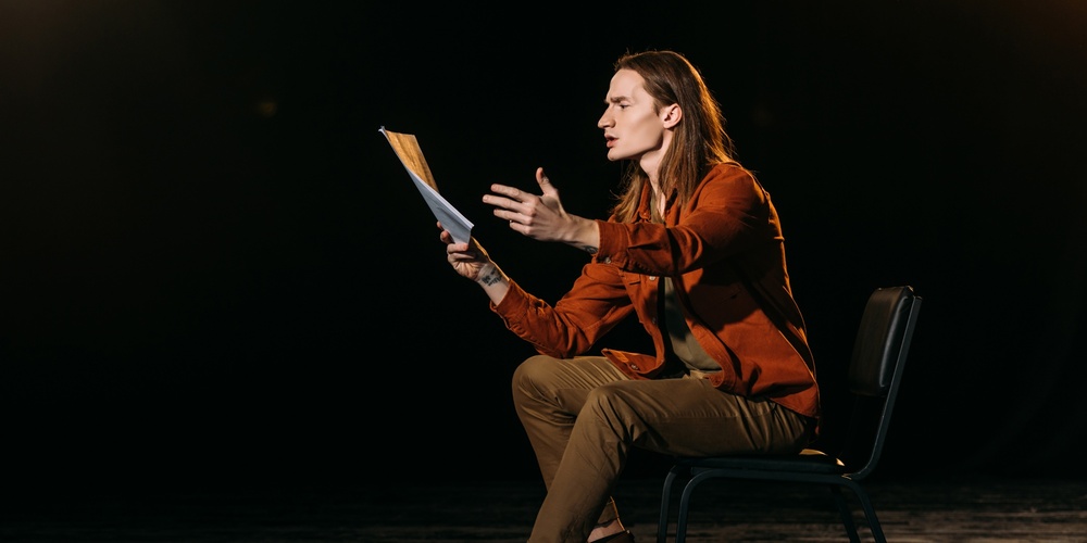 En person sitter på en stol på en scen och gestikulerar med ett manus i handen under en teaterövning.
