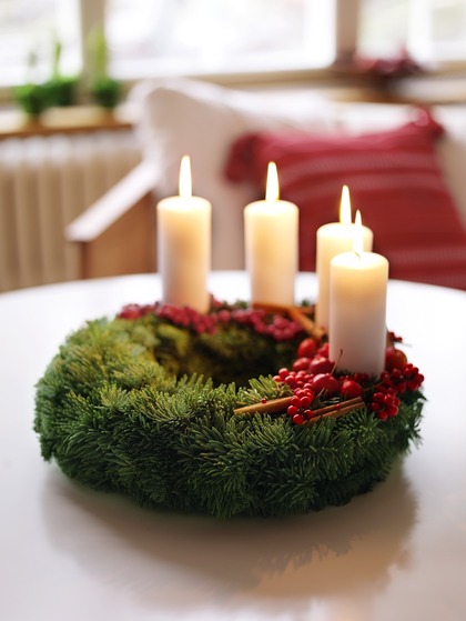 En adventskrans med fyra tända vita ljus, dekorerad med röda bär och kanelstänger, står på ett bord.