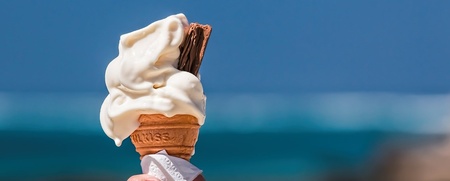 En hand håller en mjukglass i strut med chokladbit framför havet en solig dag.