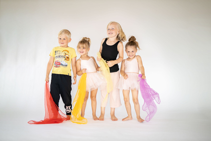 Fyra glada barn står tillsammans, tre i balettkläder och ett i t-shirt och byxor, med färgglada sjalar i handen.