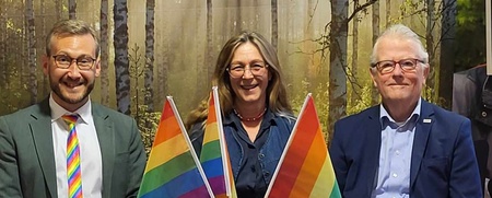 Tre personer sitter vid ett runt bord med regnbågsflaggor, i ett rum med skogstapet och affisch i bakgrunden.