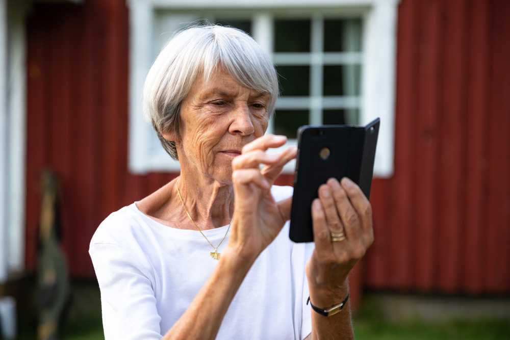 Äldre kvinna med grått hår använder en smartphone utomhus framför ett rött trähus.