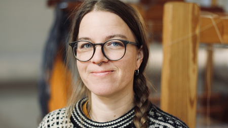 Kvinna med glasögon och fläta, bär stickad tröja, ler mot kameran inomhus.