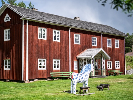 Röd trähus med vita fönster, gröna bänkar och en vitblå prickig häststaty på gräsmattan framför huset.