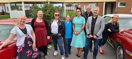 En grupp på åtta vuxna står utomhus mellan två röda bilar, klädda i olika stilar, framför ett bostadshus.