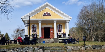 En gul byggnad med vita pelare, omgiven av människor som sitter utomhus på stolar under en klarblå himmel.
