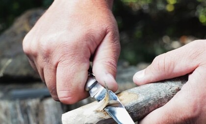 Två händer täljer en träpinne med en fällkniv utomhus.