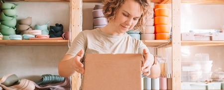 En person packar en kartong i en ljus och organiserad verkstad med hyllor fyllda av keramik och förvaringslådor.