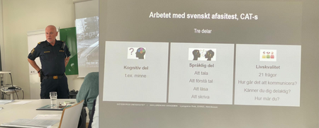 En polis står vid en projektor som visar en presentation om svenskt afasitest i ett klassrum.