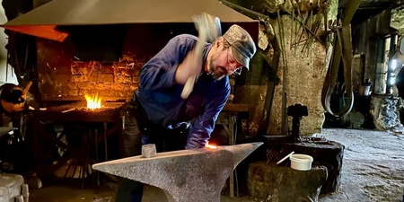 En smed arbetar vid ett städ i en traditionell smedja med eld i bakgrunden.