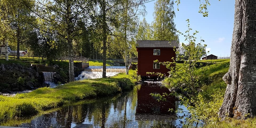 En röd stuga vid en liten fors och damm, omgiven av grönskande träd och gräs under en klarblå himmel.