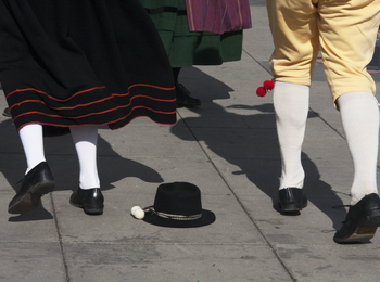 Två personer i traditionella folkdräkter dansar på en stenlagd yta, en svart hatt ligger på marken.