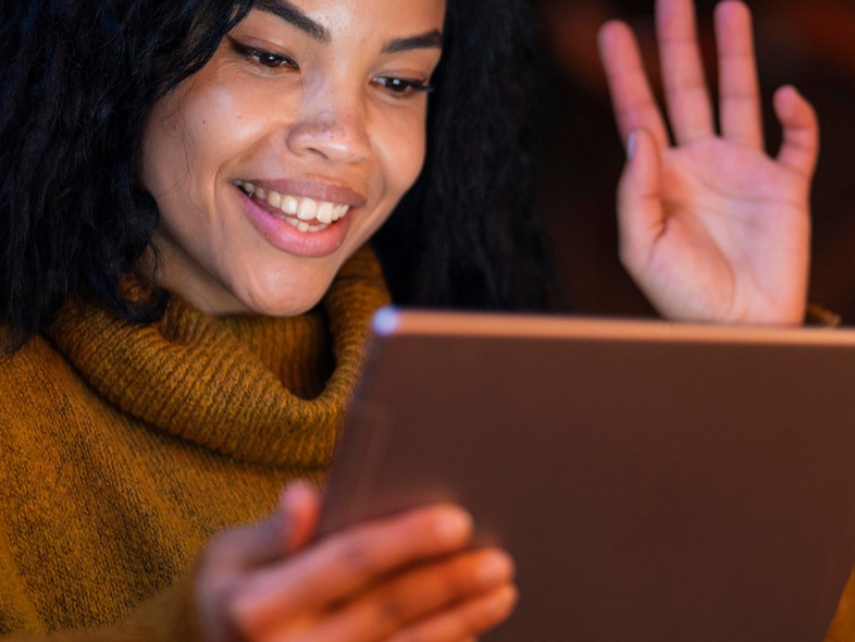 Kvinna med lockigt hår vinkar och ler mot en surfplatta i ett varmt upplyst rum.