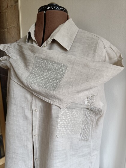 En beige skjorta med synliga lagningar och broderade lappar visas på en provdocka.