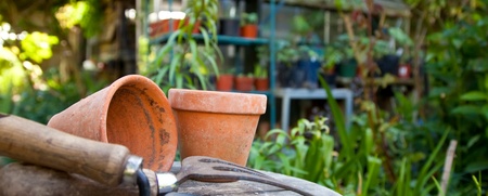 Trädgårdsredskap och lerkrukor på ett bord i en grönskande trädgård med växthus i bakgrunden.