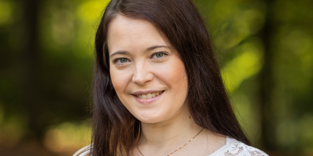 Kvinna med långt brunt hår och vit spetsblus står utomhus och ler mot kameran, grönskande bakgrund.