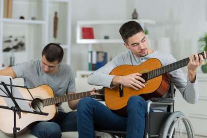 Två unga män spelar gitarr tillsammans, en sitter i rullstol och den andra läser noter.