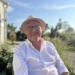 Person med vit skjorta och hatt sitter utomhus i en trädgårdsmiljö en solig dag.